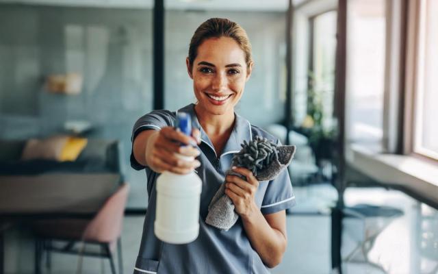 een poetsvrouw in een kantooromgeving houdt poetsproducten vast en lacht naar de camera 