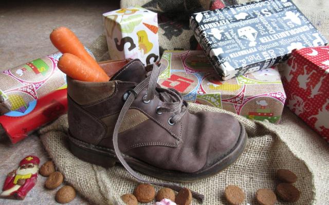 een kinderschoen met wortelen in, omgeven door pakjes en pepernoten