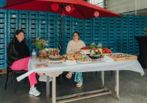 twee vrouwen die aan een tafel zitten met allemaal proevertjes rondom zich